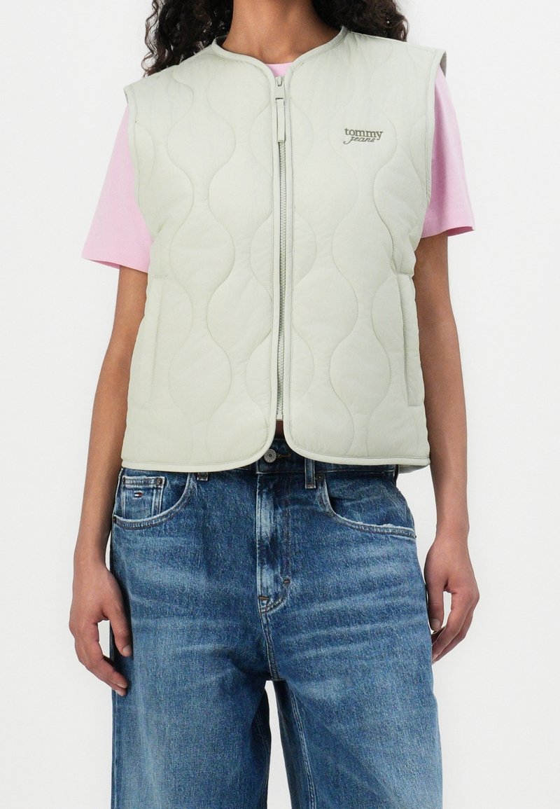 Vaaleanvihreä tikattu liivi, jossa vetoketjullinen etuosa, pyöreä pääntie ja pieni brändilogolla varustettu yksityiskohta, puettuna vaaleanpunaisen paidan päälle ja yhdistettynä sinisiin farkkuihin.