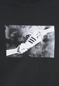 Μαύρο μπλουζάκι με μονόχρωμο γραφικό που απεικονίζει ένα χέρι να κρατά ένα παπούτσι Adidas με μαύρες ρίγες, πάνω σε υφασμένο φόντο.