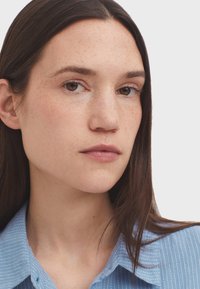 Primo piano di una persona con capelli castani di lunghezza media, che indossa una camicia a righe azzurra chiara con colletto. La pelle presenta lentiggini visibili e un aspetto naturale.