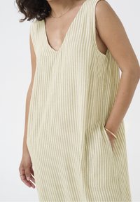 Robe d'été rayée en tissu jaune clair et blanc, avec un décolleté en V profond, sans manches et des poches latérales.