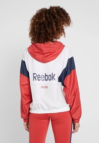 Reebok vindjacka har en vit kropp med röda och marinblå accenter, elastiska ärmslut och en justerbar huva. Logotyp tryckt på baksidan.