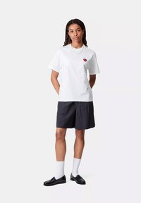 Młoda osoba z warkoczami, ubrana w białą koszulkę z czerwonym sercem, czarne teksturowane szorty, białe skarpetki i czarne mokasyny, stojąca z rękami za plecami.