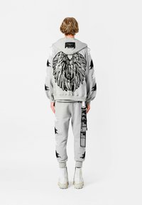 Persona con chándal gris claro con patrones de plumas en negro y blanco, gran diseño de ala plateada en la espalda y botas blancas, de pie de espaldas.