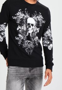 Felpa nera a maniche lunghe con un design di teschio bianco circondato da motivi floreali e ornate; polsini e orlo a coste.
