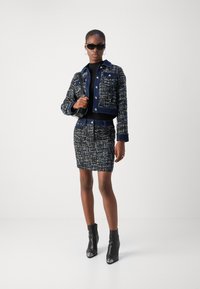 Veste en tweed navy avec une texture contrastante, des détails de boutons, une jupe assortie et des bottines noires avec des accents en argent ; le mannequin tient un petit sac.