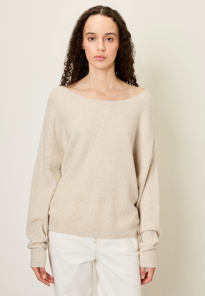 Žena s kučeravými vlasmi, oblečená v voľnom béžovom svetri spadajúcom z ramien a bielych nohaviciach, stojaca pred jednoduchým svetlým pozadím.