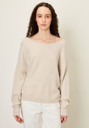 Bežový pletený sveter so širokým výstrihom a voľným strihom. Dlheé rukávy s rebrovanými manžetami. Kombinovaný s bielymi nohavicami, s hladkou textúrou.