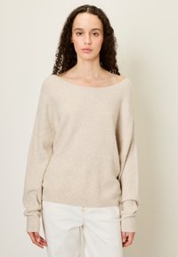 Beige gebreide trui met een brede halslijn en een loose fit. Lange mouwen met ribgebreide boorden. Gecombineerd met witte broek, met een gladde textuur.
