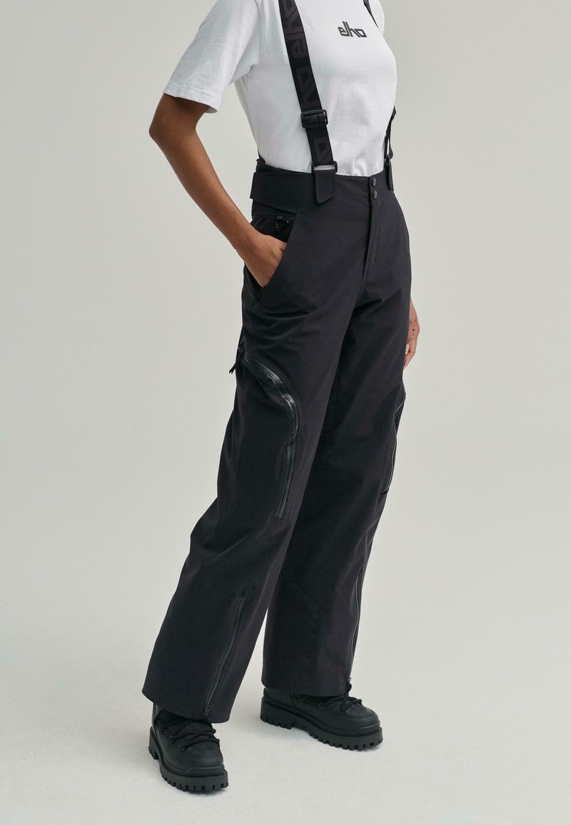 Pantaloni cargo negri cu bretele ajustabile, având buzunare laterale cu fermoar și o croială largă. Materialul are o textură netedă și un finisaj mat.