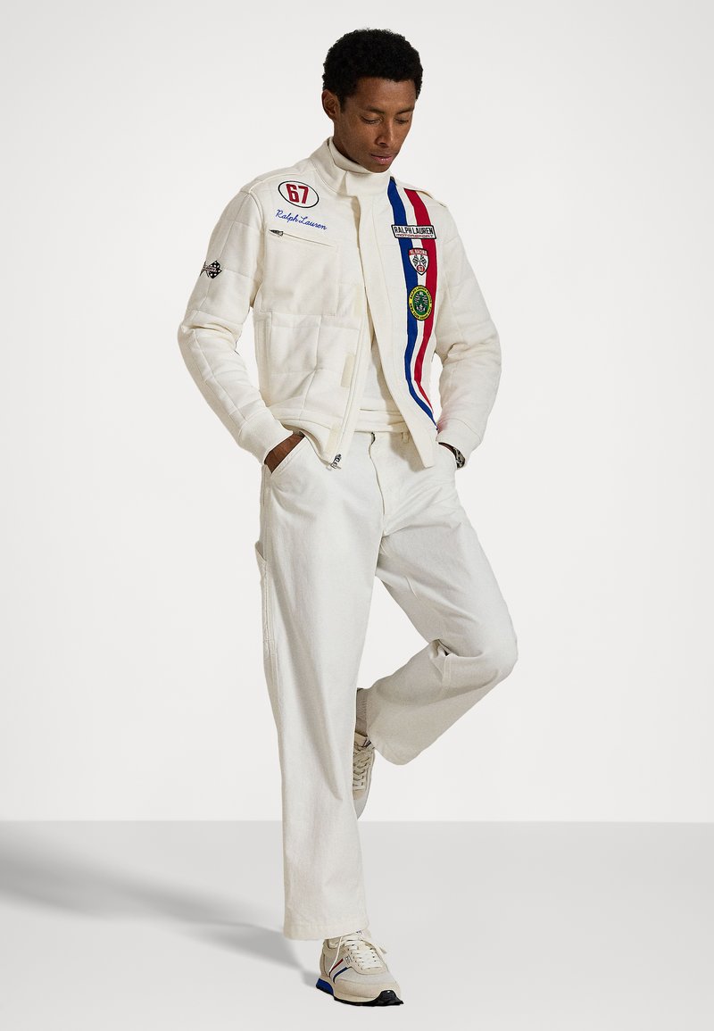 White jacket with patches and stripes, paired with white pants. Textured fabric, stand collar, and athletic shoes with blue accents.