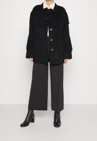 Zwarte franjejacket met een knoopsluiting, twee grote zakken en een kraag. Gepaard met donker wijde broeken en enkellaarsjes.