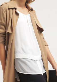 Beige Trenchcoat über einer weißen Schichtenbluse mit durchgehender Knopfleiste und offenem Kragen. Schwarze Hose vervollständigt das Outfit.