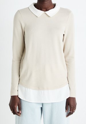 Personne portant un haut beige à manches longues avec un col blanc et un ourlet blanc superposé, associé à un jean bleu clair, debout devant un fond blanc.