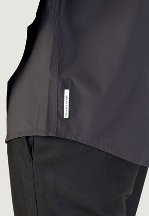 Giubbotto nero con una texture liscia e un orlo leggermente curvo, con un'etichetta con logo sul lato sinistro. Il tessuto è leggero e opaco.