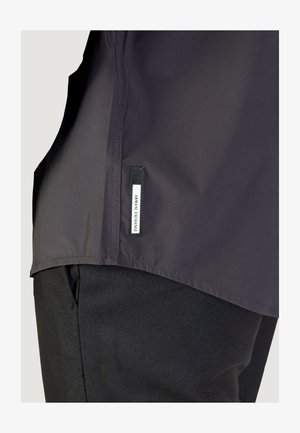 Giubbotto nero con una texture liscia e un orlo leggermente curvo, con un'etichetta con logo sul lato sinistro. Il tessuto è leggero e opaco.