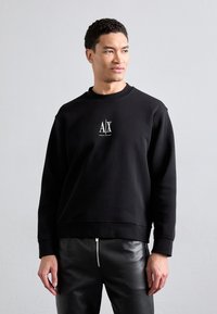 Black sweatshirt with round neckline and ribbed cuffs. Features a small white logo on the chest. Worn with fitted leather pants.
