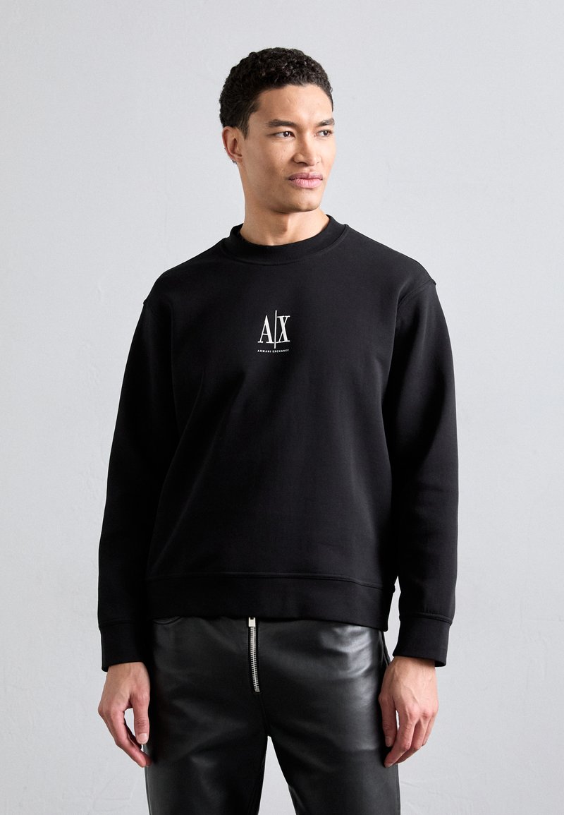Black sweatshirt with round neckline and ribbed cuffs. Features a small white logo on the chest. Worn with fitted leather pants.