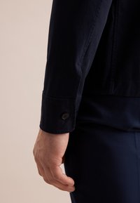 Manica di giacca blu navy dettagliata con polso a bottone, realizzata in un tessuto liscio; il braccio posizionato di lato, che rivela la camicia sottostante.