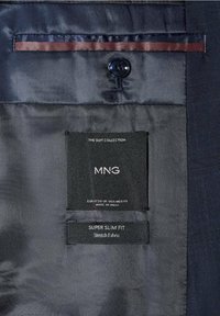 Doublure intérieure d’une veste de costume bleu marine avec bouton, comportant des étiquettes noires indiquant "MNG", "Super Slim Fit" et des détails sur le tissu/élasticité.
