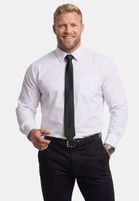 Uomo biondo sorridente che indossa una camicia bianca, cravatta nera e pantaloni, con la mano sinistra in tasca e la mano destra che gesticola in avanti.