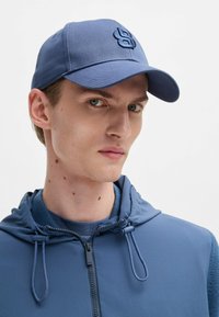 Cappello blu con superficie texturizzata, dotato di logo ricamato. Indossato con una felpa blu abbinata, con laccetti regolabili e zip.