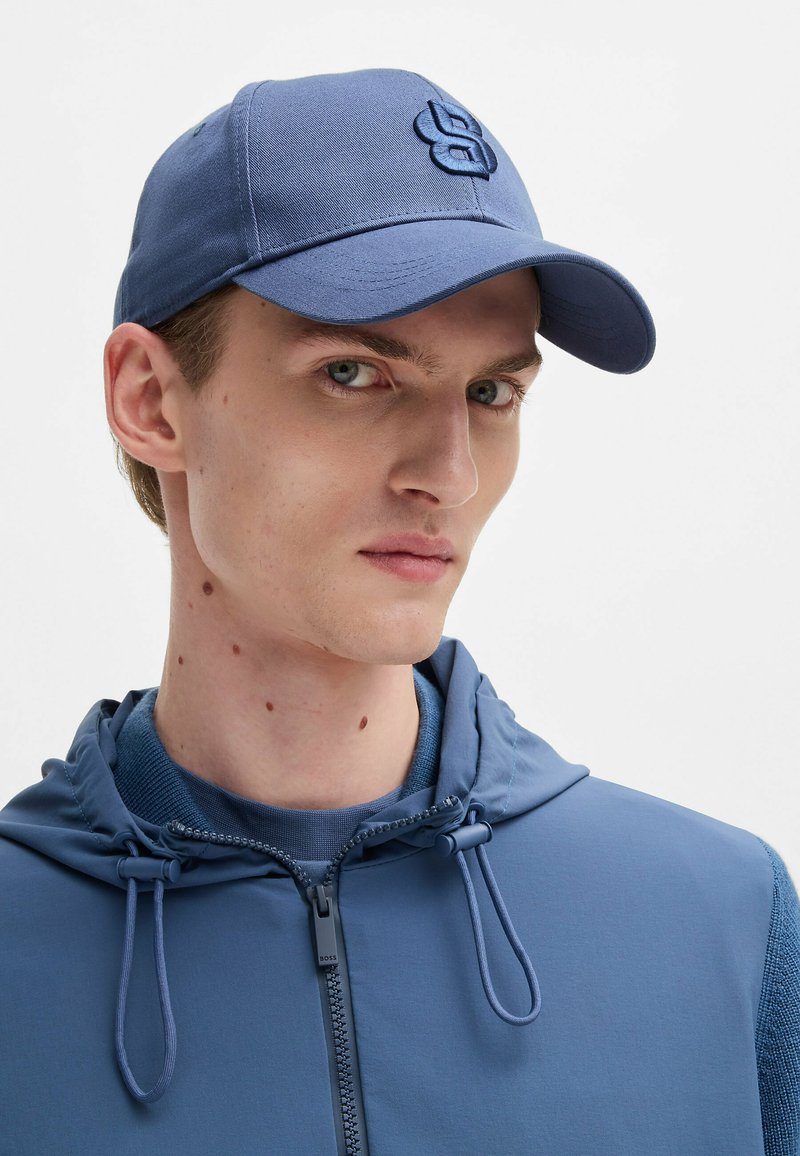 Blauwe pet met een gestructureerd oppervlak, voorzien van een geborduurd logo. Draagt een bijpassende blauwe hoodie, met verstelbare trekkoorden en een rits.