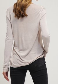 Haut beige clair à manches longues avec une coupe décontractée, caractérisé par une texture douce et un détail froncé au dos, associé à un jean foncé.