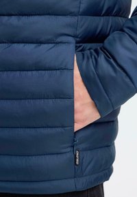 Tamno plava puffer jakna s ukrašenim stiliziranim šavovima, bočnim džepom i oznakom od tkanine. Glatka tekstura s laganim, izolirajućim dizajnom.