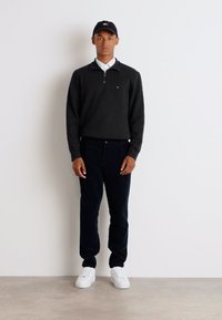 Čierny zip-up sveter s bielou košeľou pod ním, skombinovaný s tmavými nohavicami a bielymi teniskami. Model má na hlave čiernu čiapku.