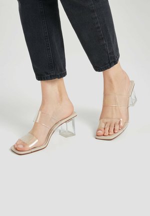 Des pieds portant des sandales à talons transparents avec des brides transparentes et des talons carrés, associés à un pantalon noir cropped sur un sol blanc.