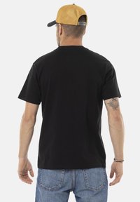 Camisa de algodón negra de manga corta, con cuello redondo, usada por una persona vista desde atrás. Acompañada de una gorra de béisbol color marrón.
