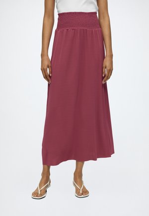 Femme portant une jupe rose foncé taille haute, longueur cheville, avec une ceinture smockée et des sandales blanches à talons.