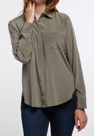 Femme portant une chemise boutonnée à manches longues vert olive avec une poche poitrine et un jean bleu foncé, main près du visage et posture détendue.