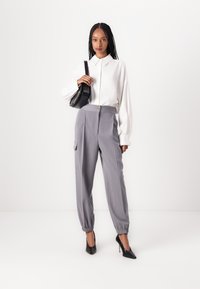 Chemise blanche à manches longues, pantalon taille haute rayé gris avec des poignets élastiques et des poches latérales, sac à main noir, et chaussures à talons hauts noires.