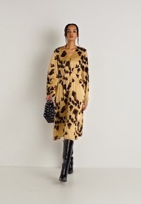 Φόρεμα με λεοπάρ τύπωμα σε μπεζ σατέν, με μακριά μανίκια, συρρυκνωμένη μέση και φαρδιά φούστα, συνδυασμένο με μια μαύρη τσάντα χειρός με λευκές λεπτομέρειες.