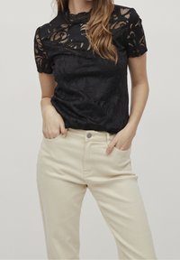 Top in pizzo nero con maniche corte, caratterizzato da un motivo floreale e un collo scallopato, abbinato a pantaloni beige chiaro a vita alta.