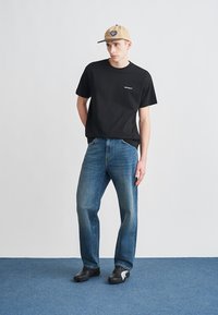 Tricou negru din bumbac cu logo alb, blugi albastri din denim și pantofi negri cu accente gri, completate de o șapcă bej cu logo.