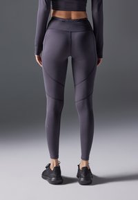 Hoge taille, aansluitende legging in donkergrijze stof met textuurpanelen en contrasterende naden, gecombineerd met zwarte sportschoenen.