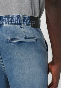 Κοντινό πλάνο ατόμου που φοράει ανοιχτό μπλε τζιν με ελαστική μέση, πίσω τσέπη και μαύρο λογότυπο, μαζί με γκρι πουκάμισο και σκουρόχρωμο δέρμα χεριού.