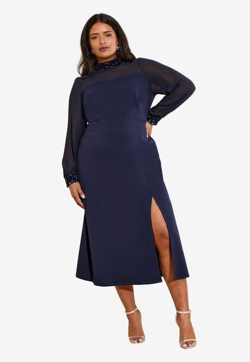 Rochie bleumarin cu mâneci lungi transparente și gât înalt. Prezintă o despicătură laterală și manșete accentuate cu paiete. Textură netedă și croi ajustat.