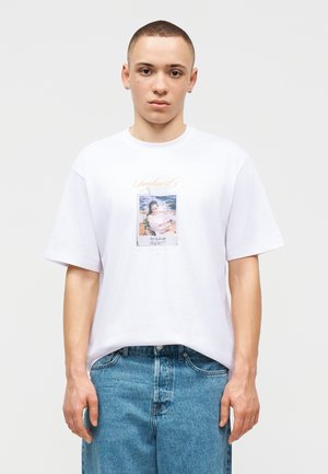 Jeune homme aux cheveux courts portant un t-shirt blanc à motifs et un jean bleu, debout devant un fond clair uni.