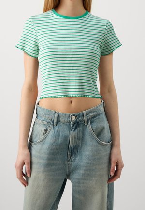 Kobieta ubrana w zielono-biały, paskowany krótki T-shirt oraz jasnoniebieskie, wysok taliowane dżinsy, stojąca z ramionami luźno opuszczonymi wzdłuż ciała.