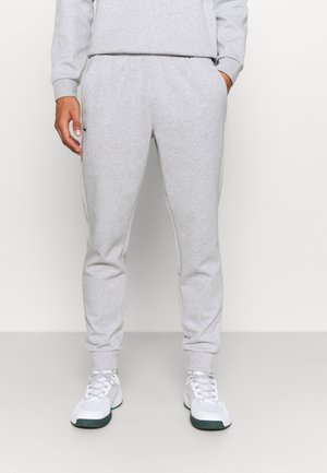 Mann trägt hellgraue Jogginghose und weiße Sportschuhe, steht vor einem schlichten weißen Hintergrund.