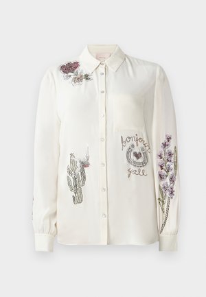 Chemise blanche à manches longues, ornée de broderies en pierres précieuses représentant des fleurs et un cactus, avec une poche poitrine du côté gauche.