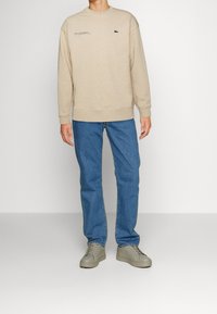 Sudadera beige con cuello redondo y logo bordado, combinada con jeans azul claro y zapatillas grises. Textura de tela suave.