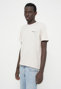 Μπεζ βαμβακερό t-shirt με κοντά μανίκια, που φέρει ένα μικρό καφέ λογότυπο με την επωνυμία στο στήθος. Φορεμένο με ανοιχτόχρωμα τζιν παντελόνια.