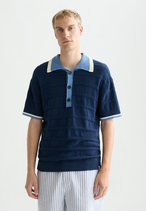 Jeune homme portant un polo bleu marine texturé à manches courtes avec des finitions bleu clair et blanches, assorti à un pantalon rayé bleu clair et blanc.