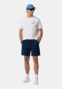 T-shirt bianca in cotone con grafica blu, pantaloni da bagno blu navy, sneakers bianche e un cappellino azzurro chiaro. Il modello sta in piedi con le mani in tasca.