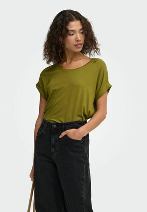 Frau mit lockigem Haar, die ein olivgrünes kurzärmeliges Oberteil und hoch taillierte schwarze Jeans trägt, mit einer Hand in der Tasche, schaut nach unten zur Seite.