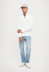 Vit zip-up sweatshirt med en logotyp, ljusblå jeans och beige sneakers. Modellen bär också en matchande beige keps.